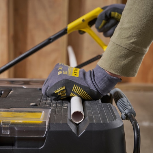 STANLEY 1-92-904 photography of gloved hand sawing PVC pipe on black workbench in workshop.
