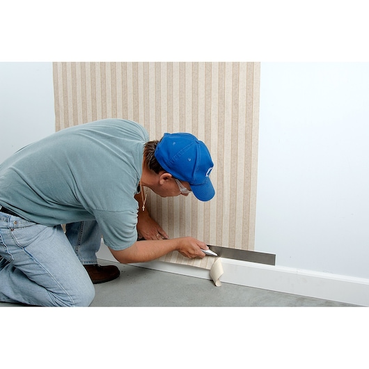 STANLEY 10-789 utility knife trimming wallpaper at baseboard, person kneeling on floor.