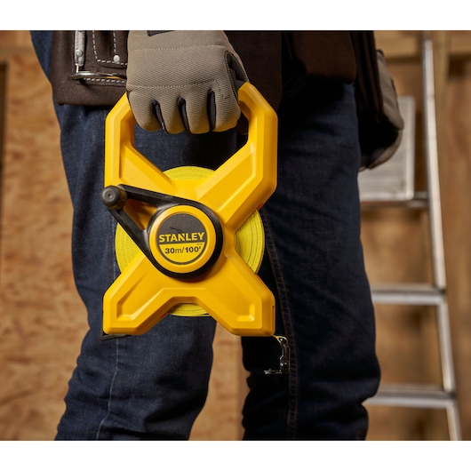 Photo of STANLEY 34-791 open reel measuring tape, yellow casing, held by a gloved hand at a construction site.