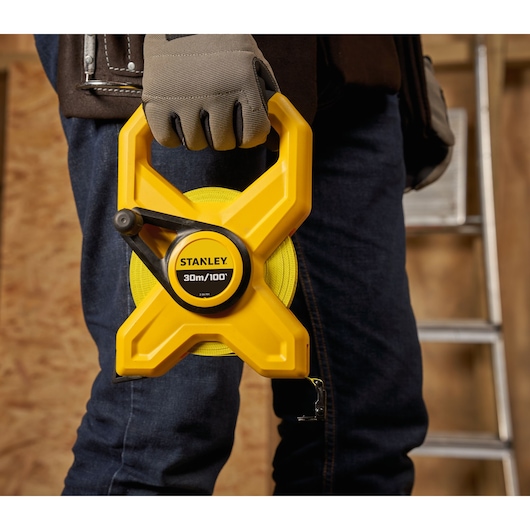 Photo of STANLEY 34-791 open reel tape measure, yellow, held by gloved hand at construction site.