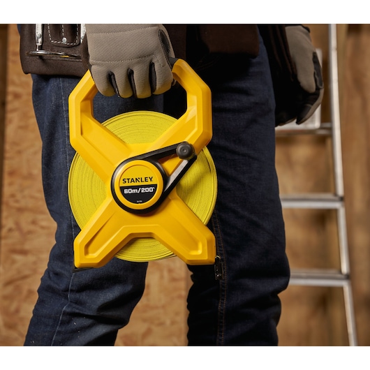 Photo of STANLEY 2-34-794 yellow open reel tape measure held by a person wearing gloves, construction background.