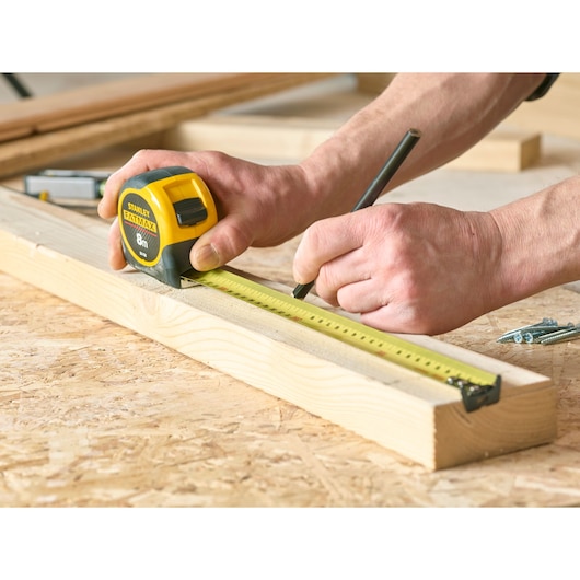 STANLEY 33-732 tape measure held against wooden plank, person marking measurements on workbench.