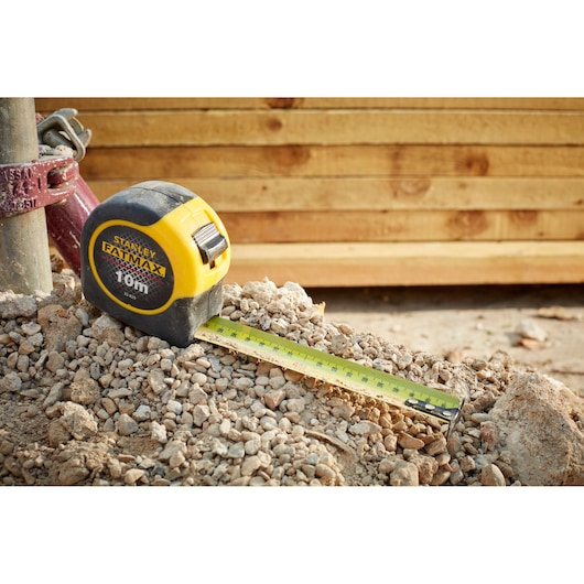 Stanley 33-829 tape measure with yellow casing on rocky ground, tape extended, wooden boards in background.