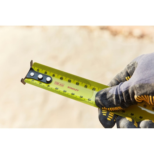Close-up photo of a STANLEY 33-829 yellow tape measure held by a gloved hand, showing measurement markings.