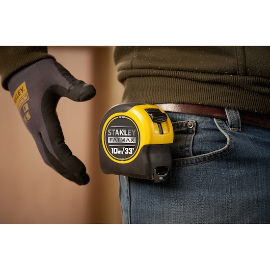 A Stanley FatMax tape measure with a 10 meter or 33 foot length is attached to a person's belt. The person is wearing jeans and a dark glove with Stanley branding.