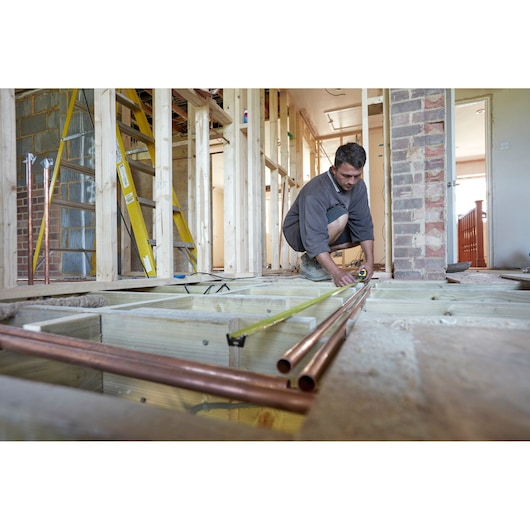 Photo of STANLEY FMHT36328 tape measure in use during home construction, measuring floor joists beside copper pipes.