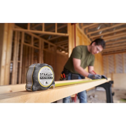 STANLEY FMHT38219-3 tape measure on wooden beam at construction site, blurred worker in background.