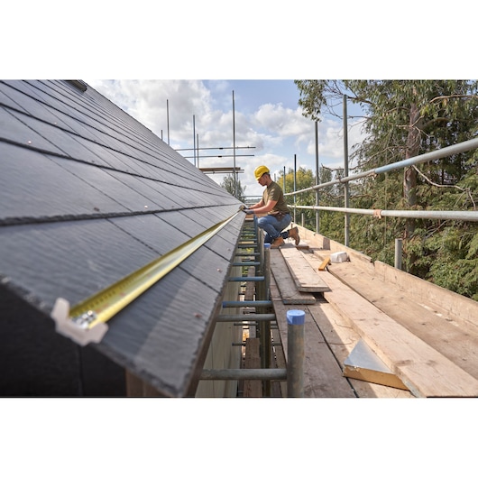 STANLEY FMHT38219-3 tape measure extended on roof next to worker wearing helmet on scaffolding.