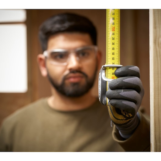 STANLEY FMHT38219-3 tape measure held vertically by gloved hand, yellow blade visible, indoor setting.