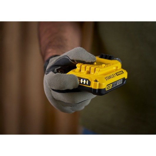 Photo of a gloved hand holding a STANLEY V20 2.0Ah battery, SKU SFMCB202, power tool accessory.