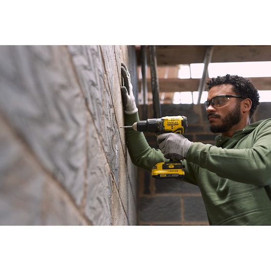 Photo of a person using a STANLEY SFMCD715D1A cordless drill on a brick wall, wearing gloves, side view.