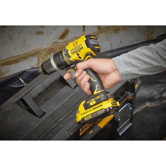 STANLEY SFMCD721DR2 cordless drill held by hand, yellow and black, near tool case, angled view.