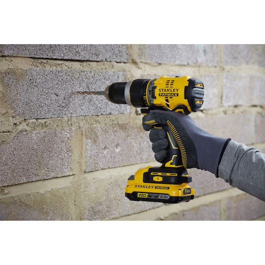 Stanley SFMCD721DR2 cordless drill held against brick wall, yellow and black design, close-up side view.