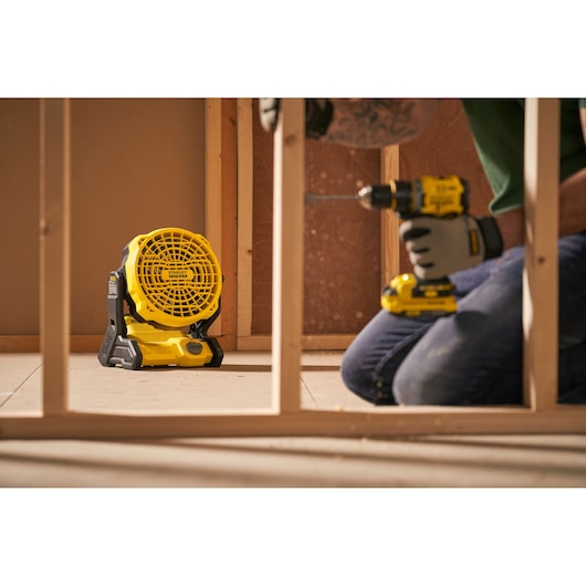 Stanley SFMCE001B portable jobsite fan on floor in construction setting, worker using power drill nearby.