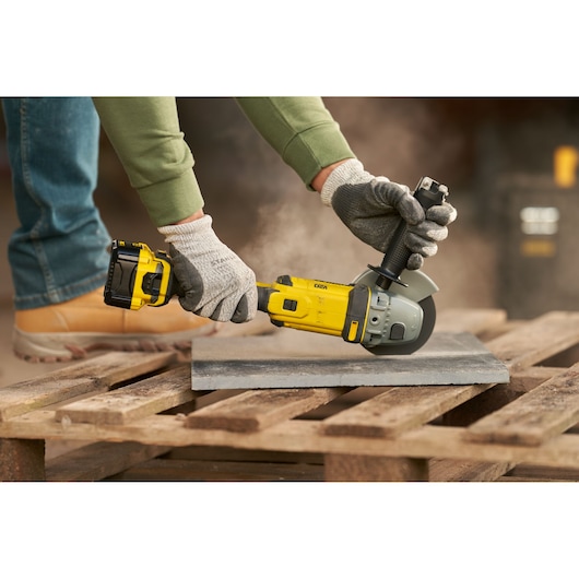 STANLEY SFMCG400M1S angle grinder in use cutting a tile, worker wearing gloves and boots outdoors.
