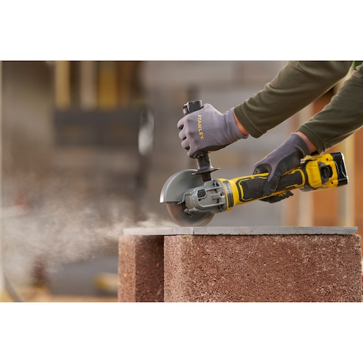 STANLEY SFMCG700B cordless angle grinder in use cutting concrete block, close-up, shows dust and gloved hands.