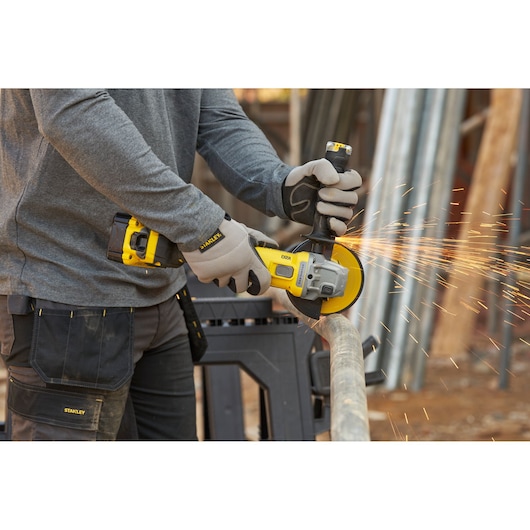 Stanley SFMCG710B-XJ cordless angle grinder in use cutting metal pipe, yellow and black tool, sparks visible.