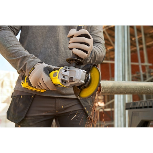 Photo of worker using STANLEY SFMCG710B-XJ cordless angle grinder cutting metal pipe, sparks flying, workshop setting.