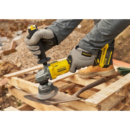 Stanley SFMCG710B-XJ cordless grinder in use, yellow and black, shown polishing a shovel blade outdoors.