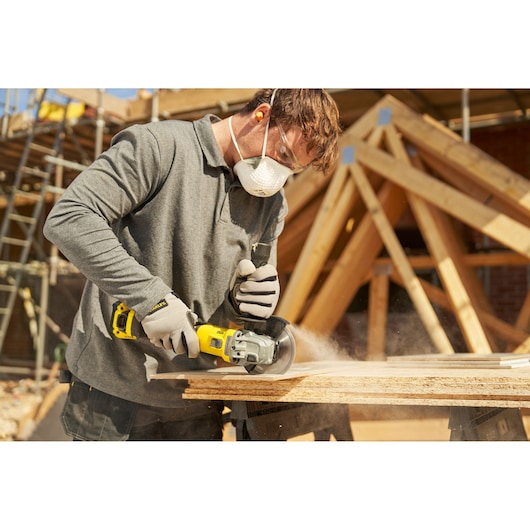 STANLEY SFMCG710B-XJ angle grinder in use cutting wood at a construction site, worker wearing gloves and mask.