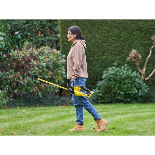 STANLEY SFMCHT650D1 hedge trimmer in use outdoors, person holding yellow and black tool near green bushes.