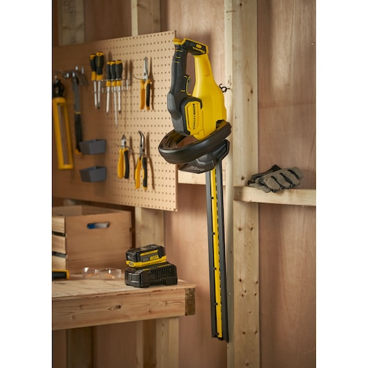 STANLEY SFMCHT655M1 cordless hedge trimmer hanging in workshop with battery and charger on table.