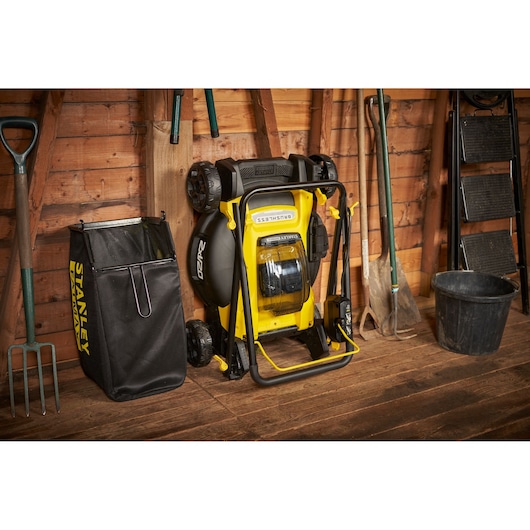 STANLEY SFMCMW2651M lawn mower shown folded upright next to black grass collection bag in a wooden shed.