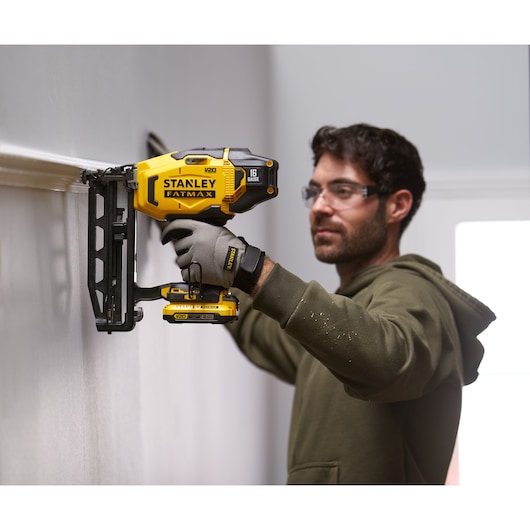 Stanley SFMCN616B cordless finish nailer in use, yellow and black, held against a wall by a person wearing gloves.