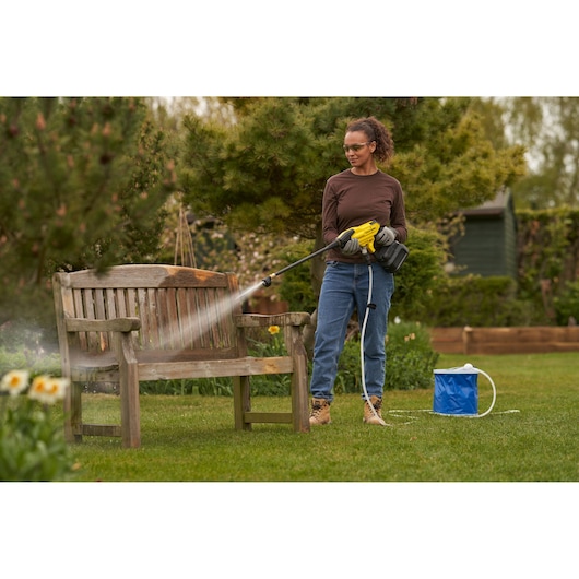 STANLEY SFMCPC93B portable pressure washer spraying wooden bench outdoors, with blue water container on grass.