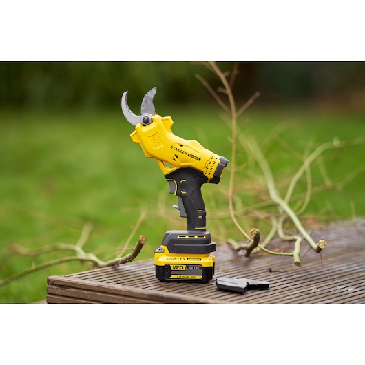 STANLEY SFMCPP32B cordless pruning shears on table outdoors with branches and battery visible.