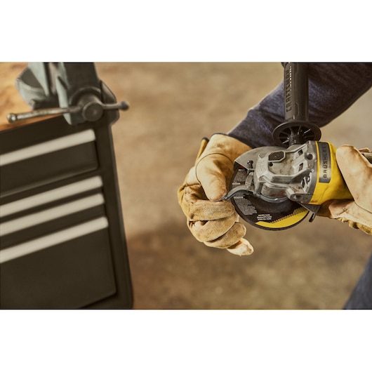 STANLEY SFMCS310B cordless angle grinder held by gloved hands near workshop cabinet.
