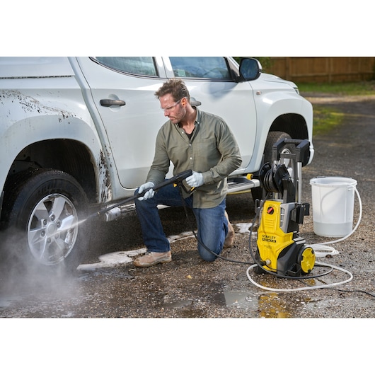 Stanley SFMPW2400 pressure washer cleaning car wheel outdoors; yellow unit, hose, bucket in scene.
