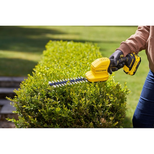 STANLEY STCSS20B cordless hedge trimmer in use trimming green bushes outdoors.