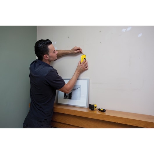 STANLEY STHT0-77403 stud finder in use on wall, yellow handheld device, photo of product in home setting.
