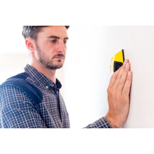 STANLEY STHT0-77404 yellow stud finder held against a wall by a person wearing a checkered shirt.