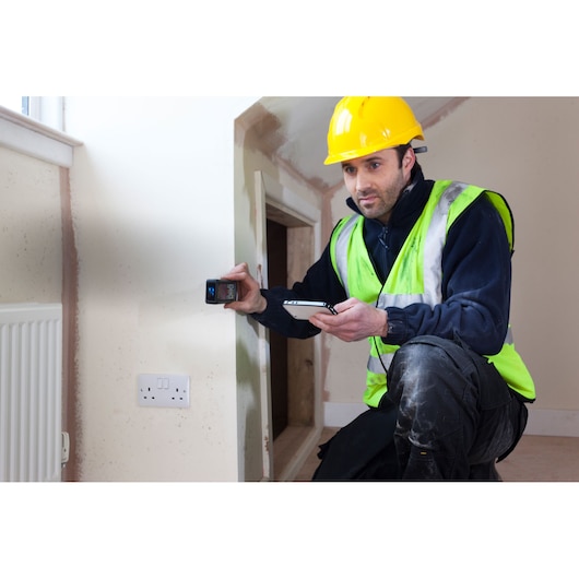 Photo of worker using STANLEY STHT1-77347 laser distance measurer in home, wearing safety vest and helmet.