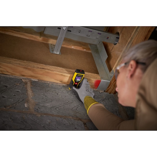 STANLEY STHT1-77409 laser distance measurer held by gloved hand measuring wood beam indoors.