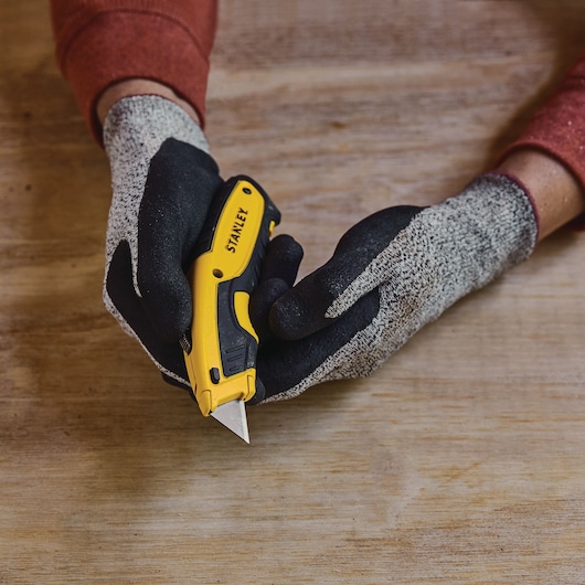Photo of STANLEY STHT10479-0 utility knife held in gloved hands, yellow and black, on wood surface.