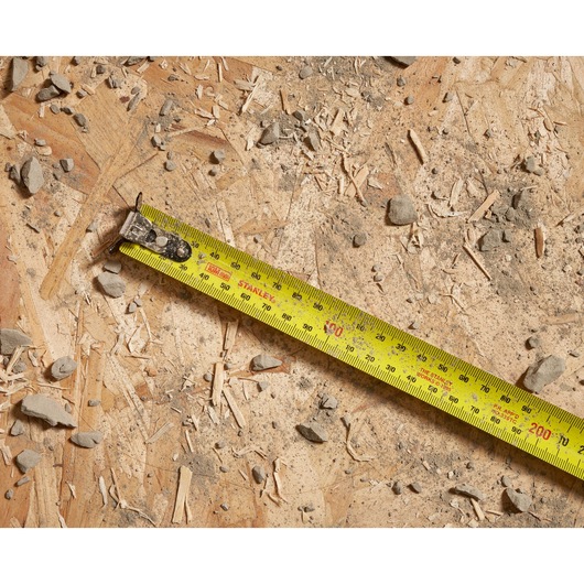 Photo of STANLEY STHT37241 tape measure extended on rough plywood with debris, showing metric and imperial markings.