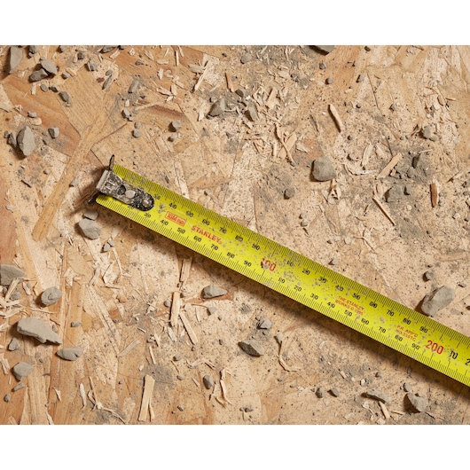Photo of STANLEY STHT37241 tape measure extended on rough plywood with debris, showing metric and imperial markings.