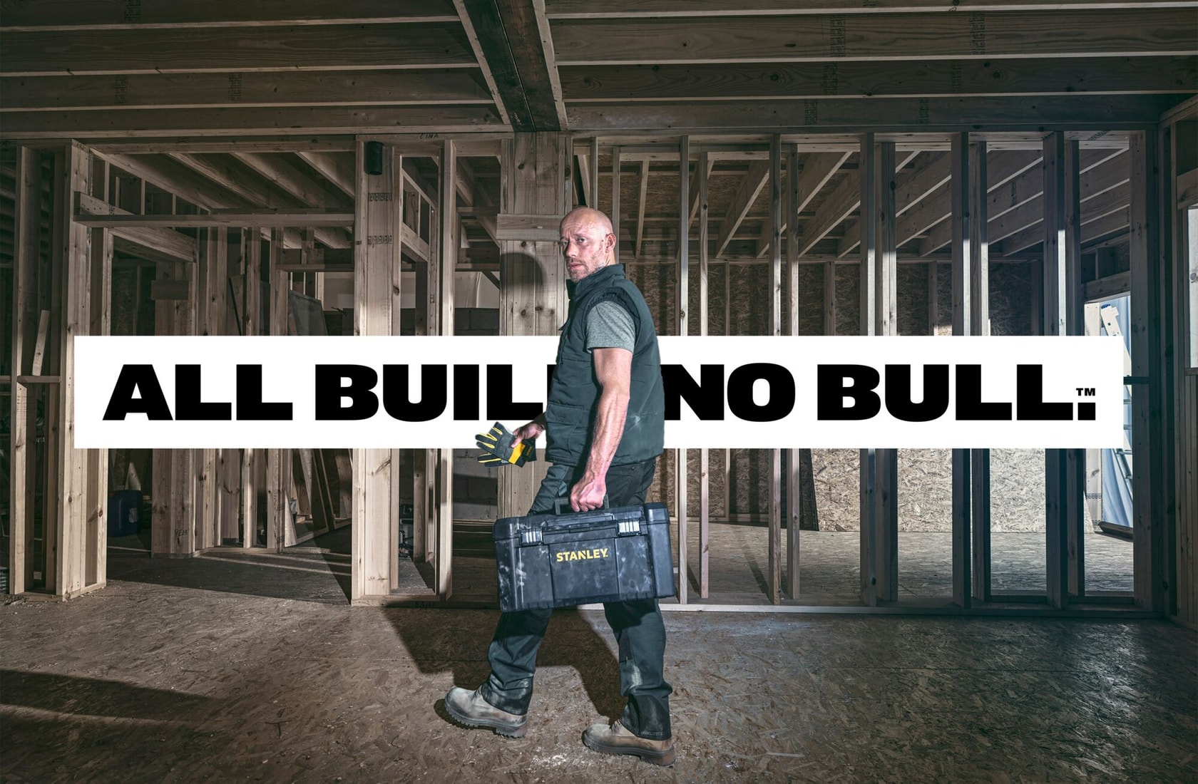 STANLEY toolbox carried by worker in unfinished wood frame house, text ALL BUILD. NO BULL. across center.